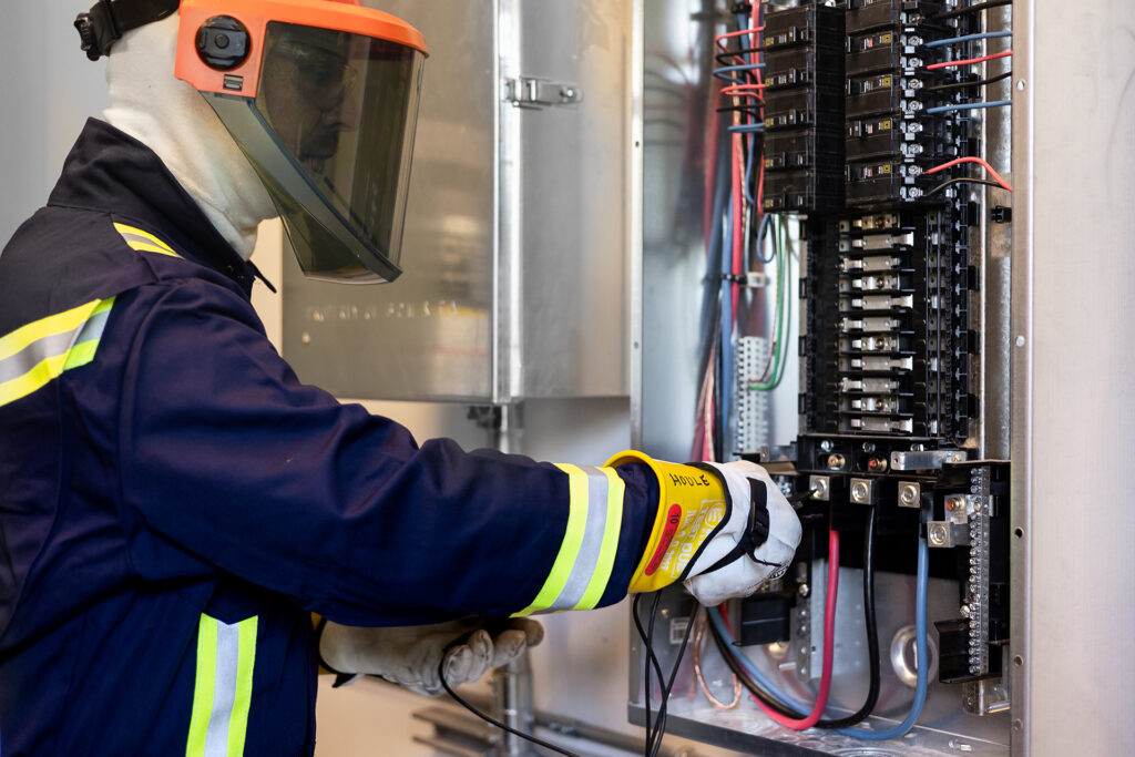 The image shows an individual wearing a high-visibility jacket with reflective stripes, safety gloves, and a face shield, working on an open electrical panel. The panel reveals multiple wires and electrical components, indicating that the person is inspecting or repairing it. This highlights the importance of safety measures when dealing with electrical systems.